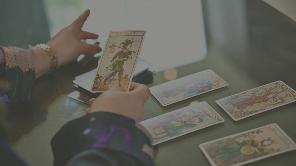 Jeu de tarot posé sur la table, avec une carte en évidence dans les mains d’Inès Rossignol représentant la carte du Mat, figure symbolisant la liberté, l'inconnu et les nouveaux départs.
