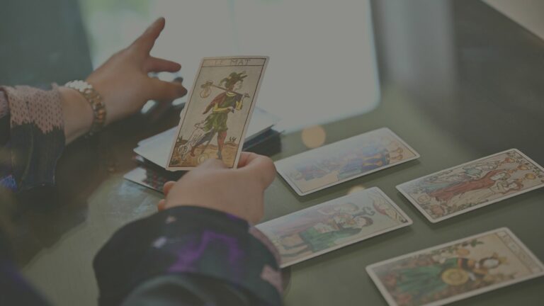 Jeu de tarot posé sur la table, avec une carte en évidence dans les mains d’Inès Rossignol représentant la carte du Mat, figure symbolisant la liberté, l'inconnu et les nouveaux départs.